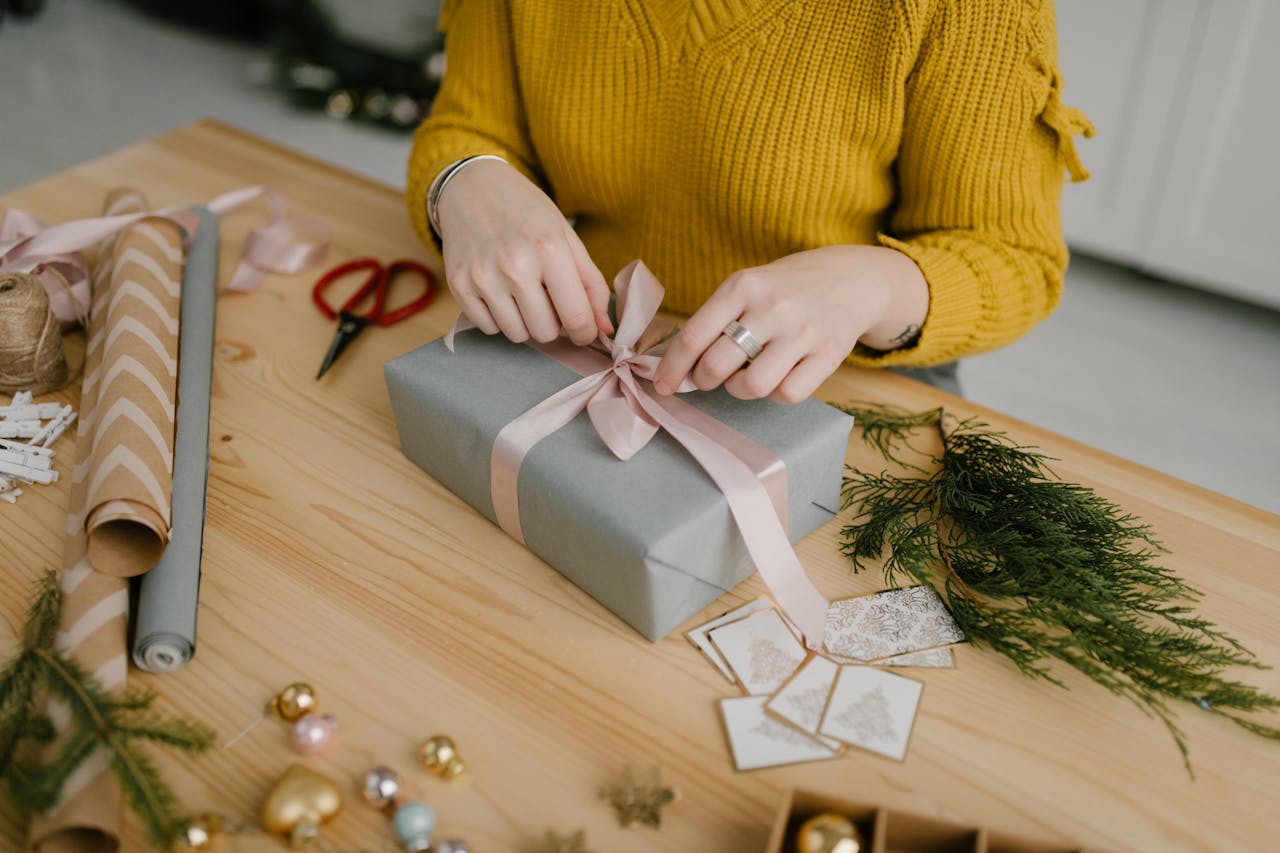 hero-img-01 Woman in yellow sweater wrapping a gift with pink ribbon on wooden table decorated for Christmas.