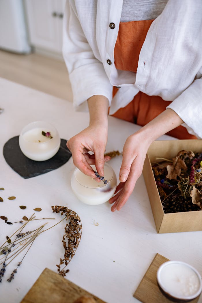 gallery-3 Hands crafting candles with dried flowers in an artisanal workshop setting.